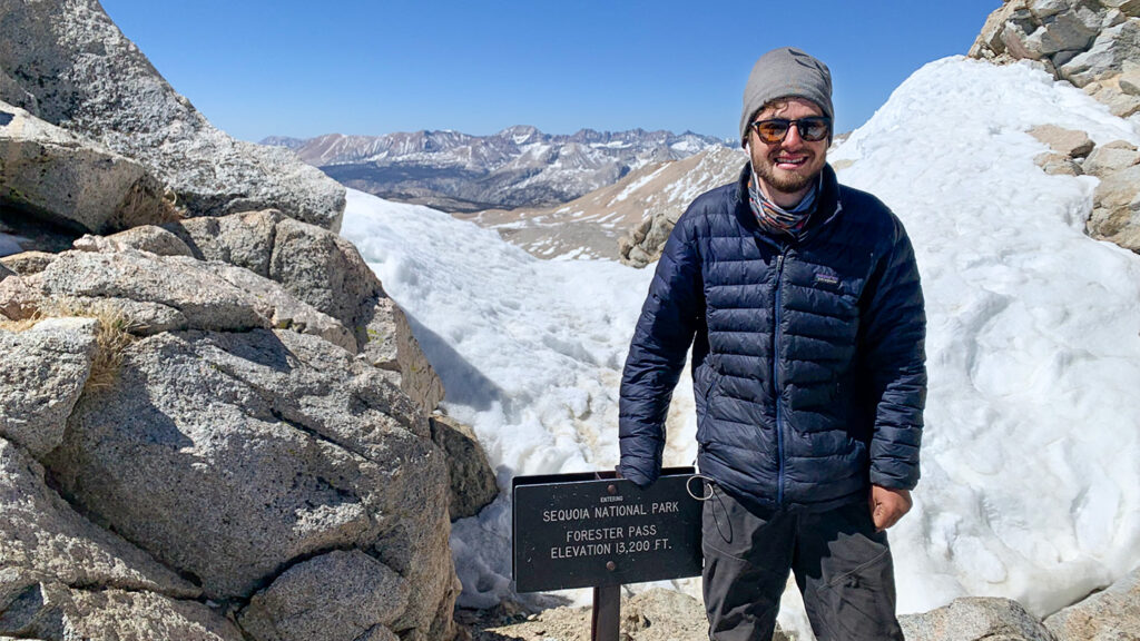 Ben Shmaruk in a winter hat and coat standing at a trail sign with snow-capped mountains behind him.