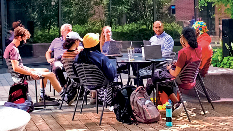 Yusuf Dahl sits with students at a table at an outdoor courtyard, laptops on table