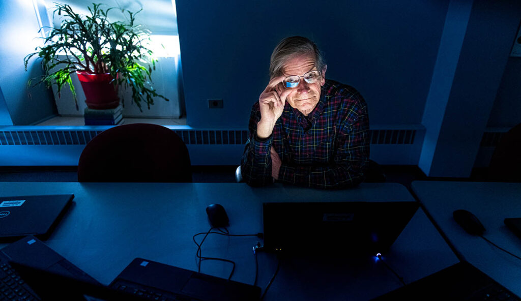 Tom Hill sits at a desk, a plant to his left, blue filtered light shines in the room
