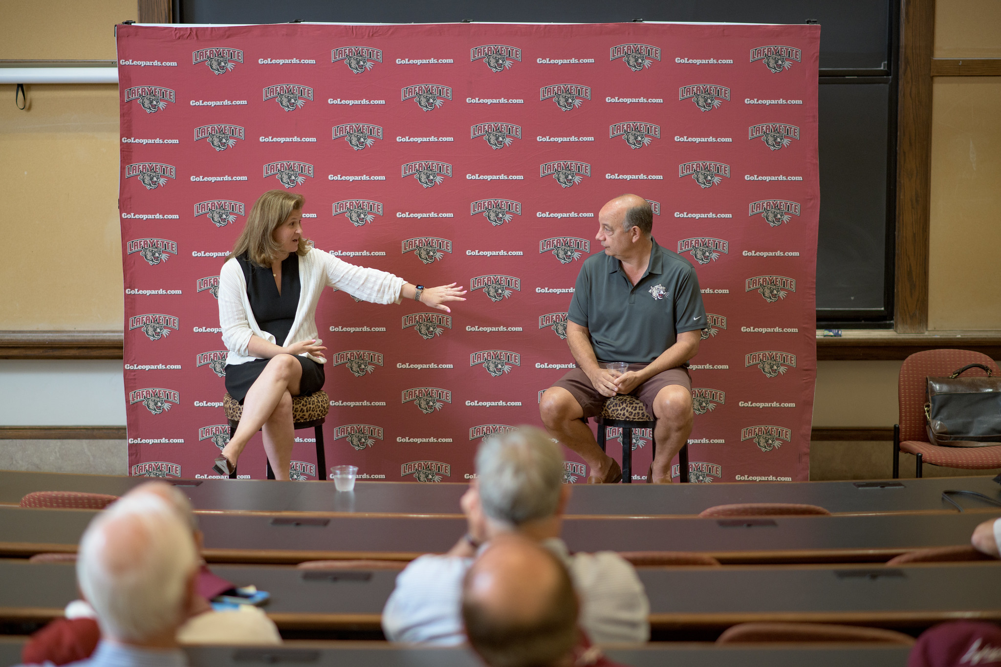 Nicole Farmer Hurd sits on a stool next to Bob Sell and talk, a Lafayette backdrop is behind them in a classroom where alumni are gathered
