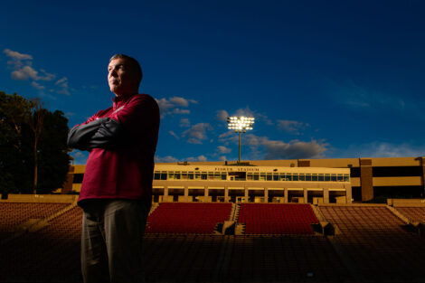 Coach John Troxell stands with arms crossed on Fisher Field, the sky behind him a deep blue