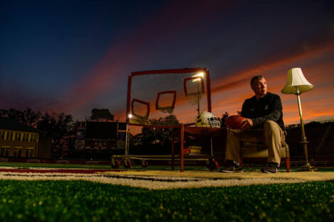 John Troxell sits on a chair with a table to his right and a lamp to his left as the sun sets on Fisher Field