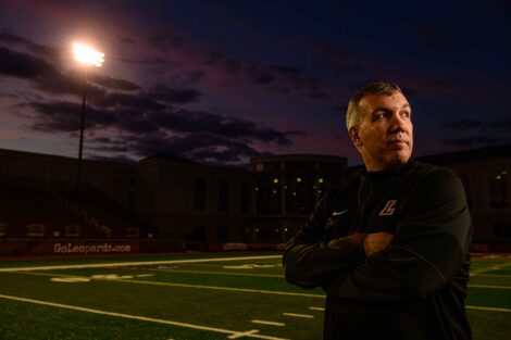 Coach John Troxell stands with arms crossed on Fisher Field, a field light illuminates behind him