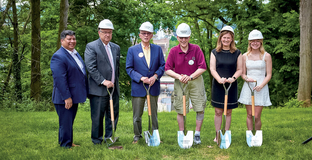 Mayor Sal Panto and President Nicole Farmer Hurd stand with others holding shovels and wearing hard hats on a grassy area of campus that overlooks Easton